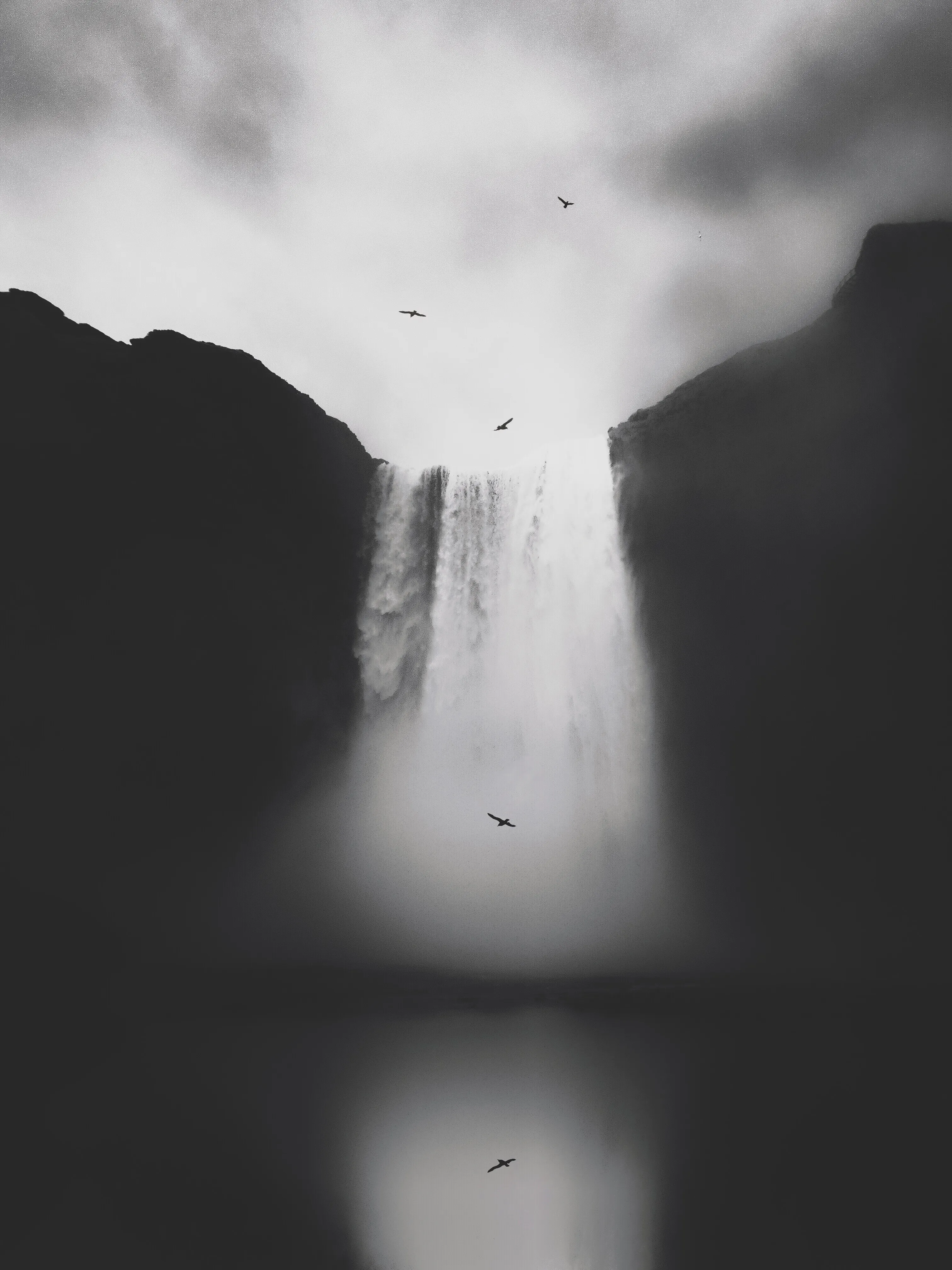 Dramatic black and white tall waterfall between dark cliffs with birds across a cloudy sky
