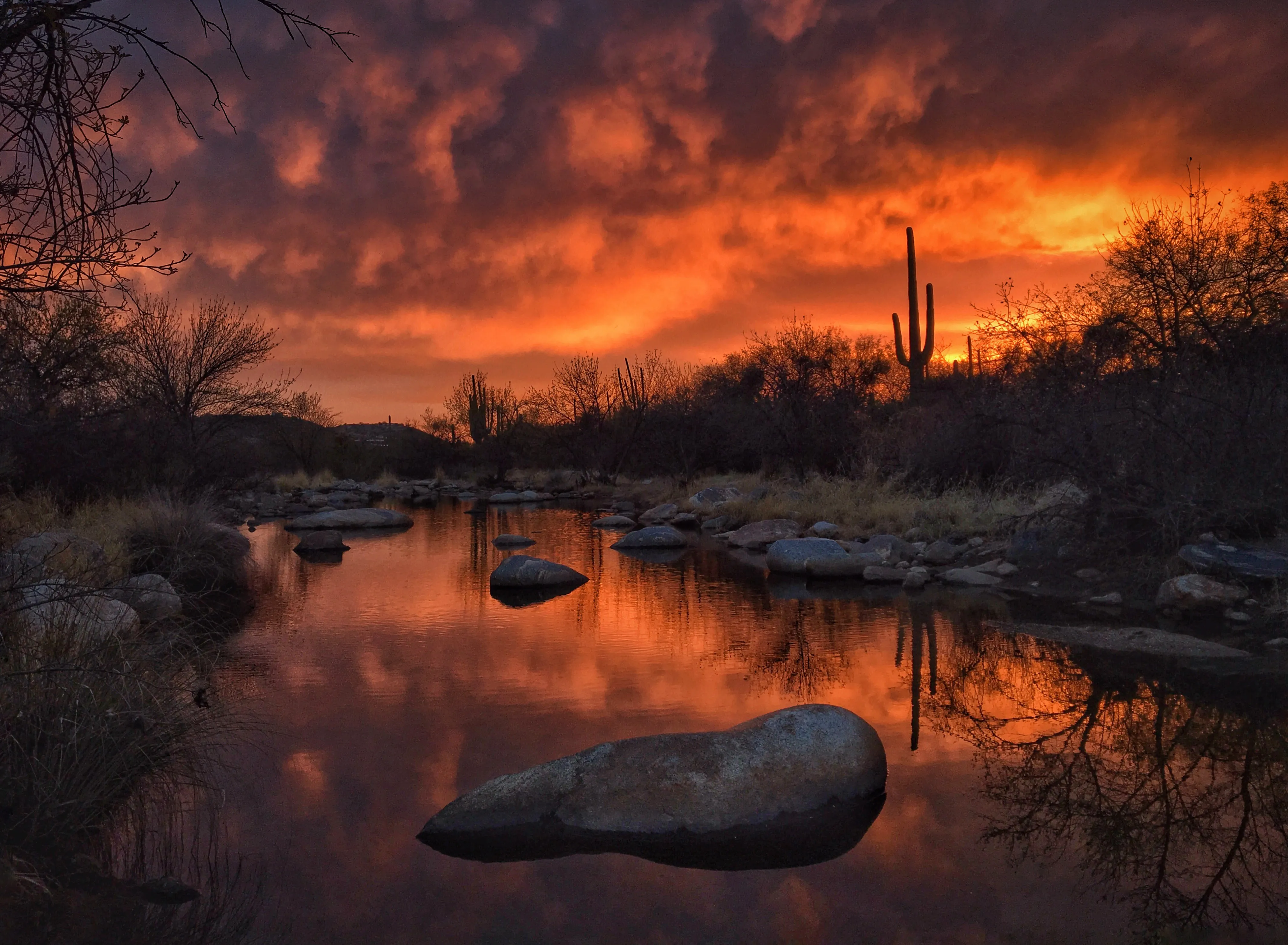 Abstract fiery orange sunset sky reflected over a calm desert stream with rocky banks, warm dramatic mood